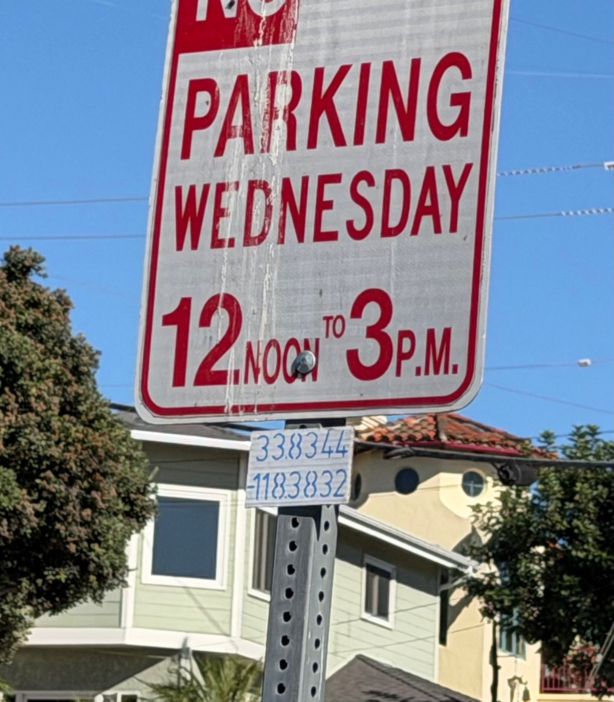 A NO PARKING WEDNESDAY 12 NOON TO 3 P.M. sign. Below it on the post, a small hand-lettered tag reads 33.8344 and -118.3832.